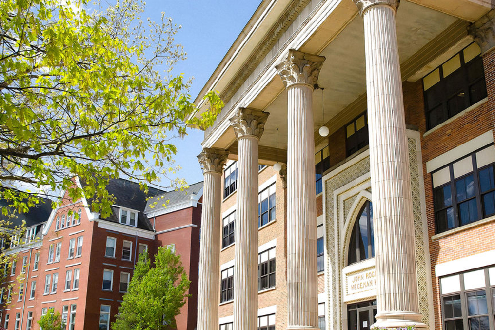 Close-up view of a historic brick academic building with tall white classical columns and decorative architectural details at the entrance. Sunlight filters through green tree branches in the foreground, with additional campus buildings visible in the background.