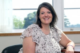 A smiling woman with shoulder-length dark hair sits indoors by a window, wearing a light floral blouse, with natural light illuminating her face.